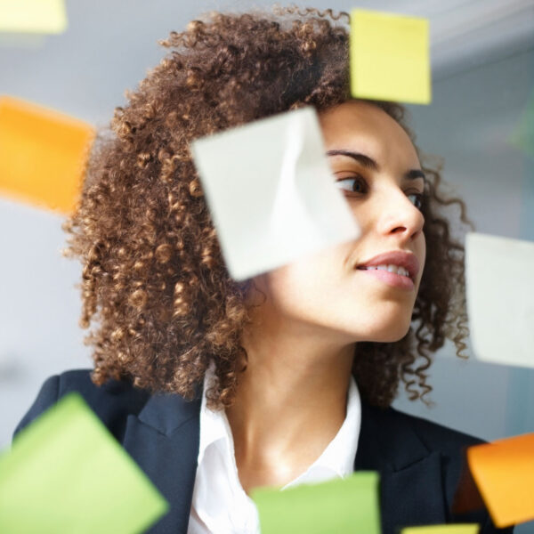 Changitors_Start_Zeit_Ort Frau mit lockigem Haar blickt konzentriert auf eine Glaswand mit bunten Klebezetteln während eines Workshops